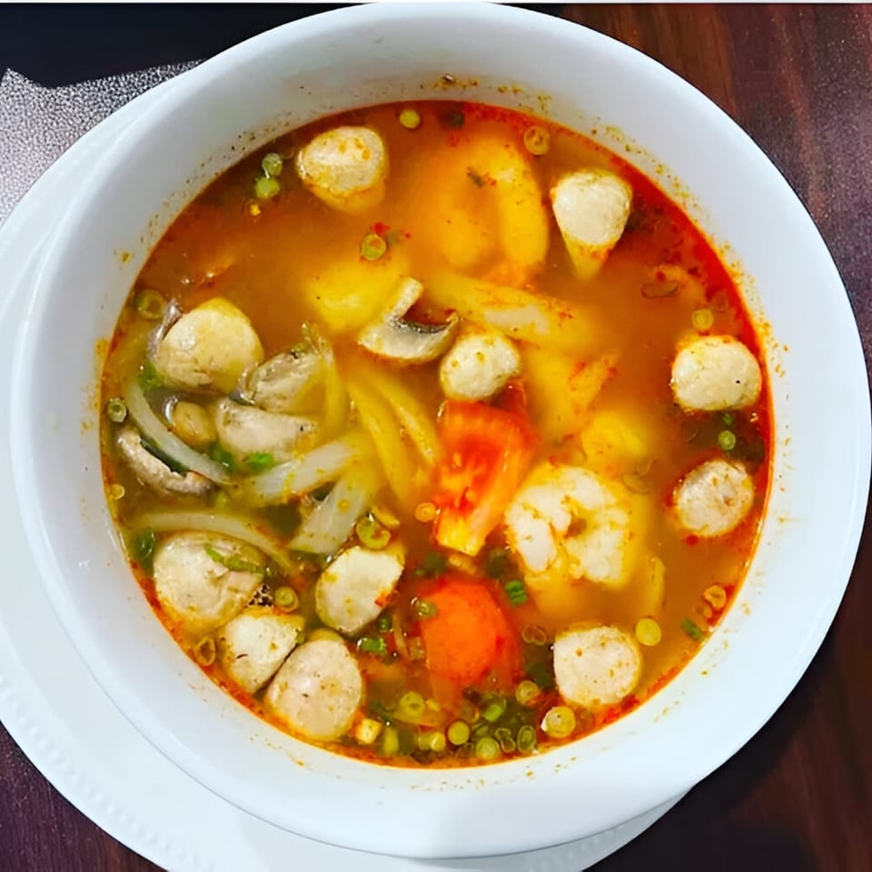 A vibrant Thai Tom Yum soup, brimming with shrimp, fish balls, mushrooms, and vegetables in a white bowl.